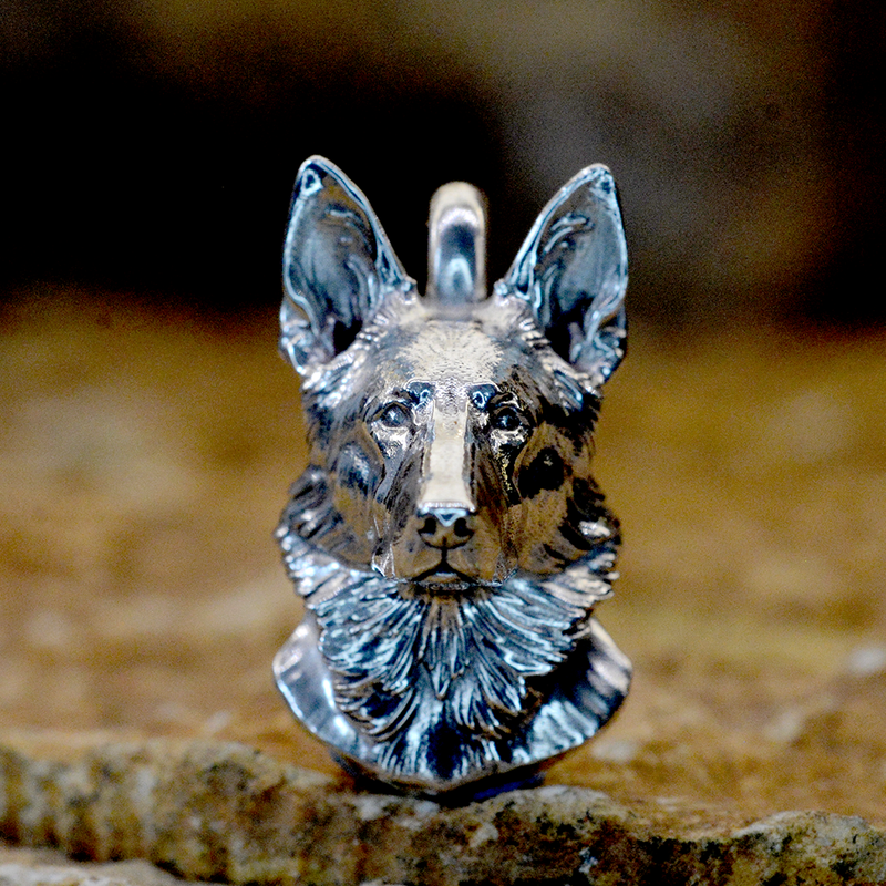 A detailed German shepherd sterling silver dog ashes pendant with a textured, metallic finish, standing on a rustic stone surface against a dark, blurred background. Front view