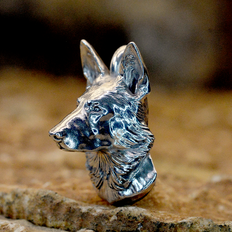 A detailed German shepherd sterling silver dog ashes pendant with a textured, metallic finish, standing on a rustic stone surface against a dark, blurred background.