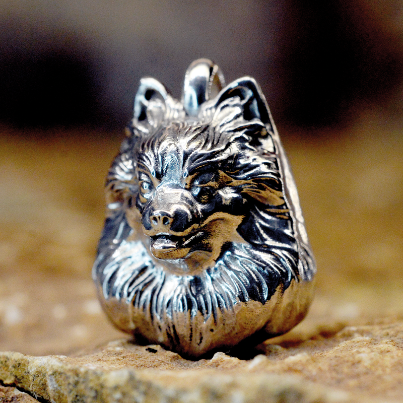 A detailed sterling silver pendant shaped like a dog's head, featuring textured fur and a polished finish, resting on a sandy surface.