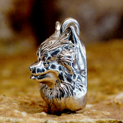 A detailed sterling silver pendant shaped like a dog's head with textured fur, shown standing on a sandy surface with a blurred brown background.