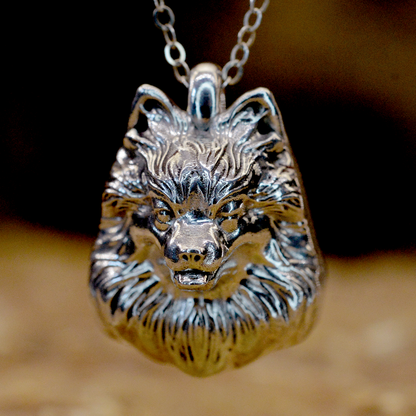 A close-up of a silver wolf pendant necklace with a detailed metallic wolf head charm on a chain, set against a blurred warm brown background.